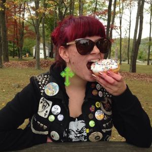 Red headed zinester wearing a black vest covered in patches and buttons eats a donut in front of trees.